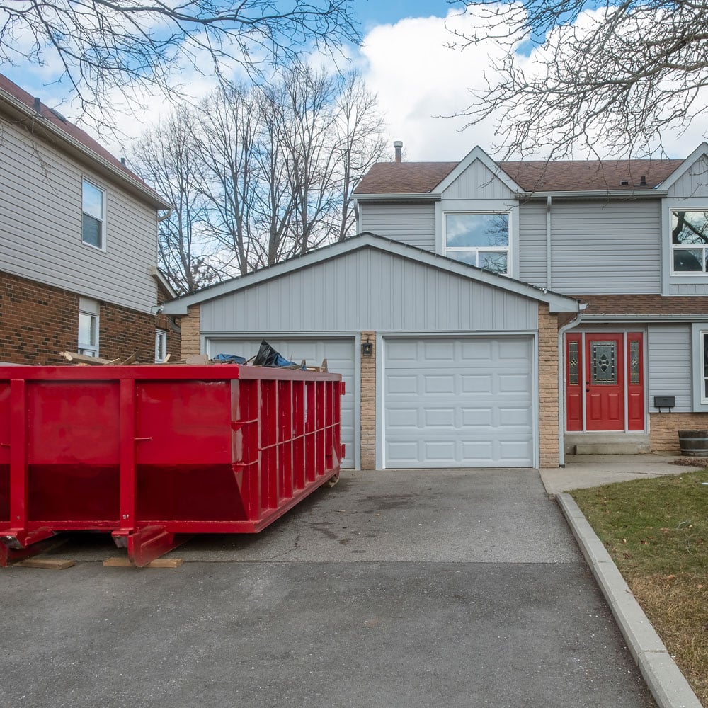 A large red dumpster sits on a driveway in front of a gray house with a two-car garage. The house features a red front door.
