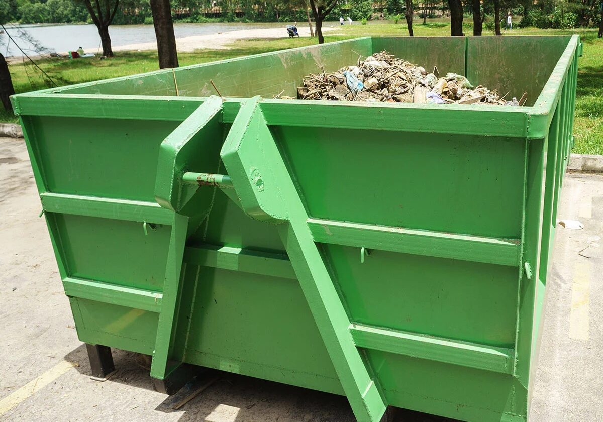 A green metal dumpster filled with construction waste. It is placed on a concrete surface.