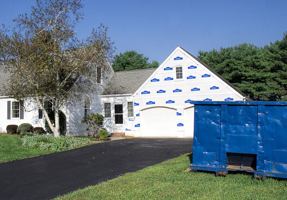 A house with blue tape on the exterior walls, a large blue dumpster, and a newly paved black driveway.